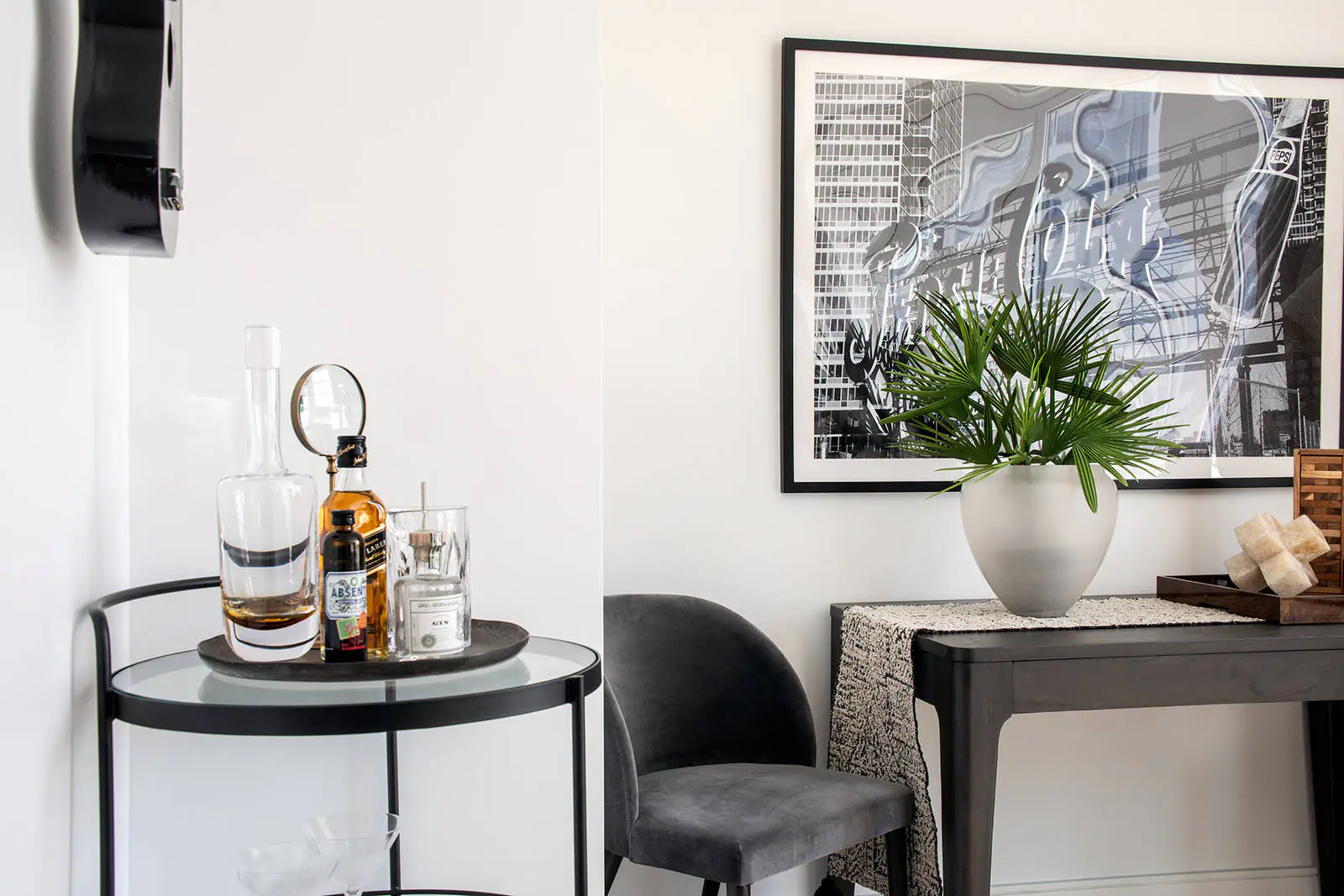 Studio bar table detail with glass bar table and drinks, dark wood entry table with dark grey velour side char, vase with plant, white walls and b&w framed photo above